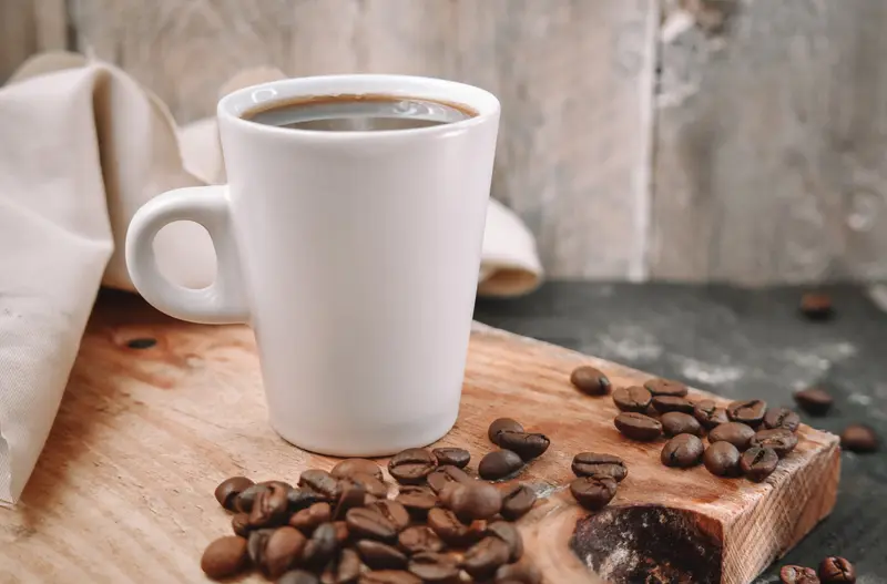 كوب أبيض فيه قهوة وحبوب بن على لوح خشبي